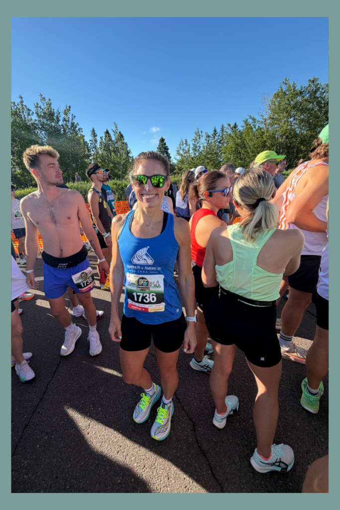 Stephanie Thomas, a licensed therapist and anxiety specialist, in a blue tank top and sunglasses standing calmly in a crowded starting corral at Grandma's Marathon, using mental training to regulate race day performance anxiety.